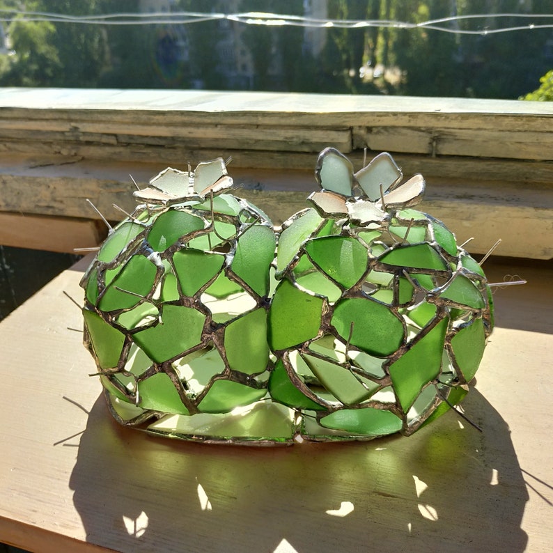 May include: A green glass cactus sculpture made of many small pieces of glass. The cactus has two parts and is sitting on a wooden surface.