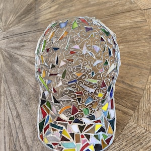 May include: A mosaic baseball cap with a wire frame and colorful glass pieces. The cap features a curved brim and a rounded crown, with a variety of shapes and colors including red, green, yellow, and blue. The cap is displayed on a wooden surface.