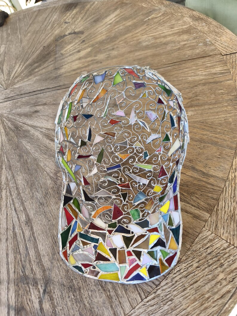 May include: A mosaic baseball cap with a wire frame and colorful glass pieces. The cap features a curved brim and a rounded crown, with a variety of shapes and colors including red, green, yellow, and blue. The cap is displayed on a wooden surface.