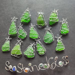 May include: Twelve green glass Christmas tree ornaments wrapped in silver wire. The ornaments are arranged in a semi-circle on a gray surface. The text "Sea Stained Glass" is written in silver wire below the ornaments.