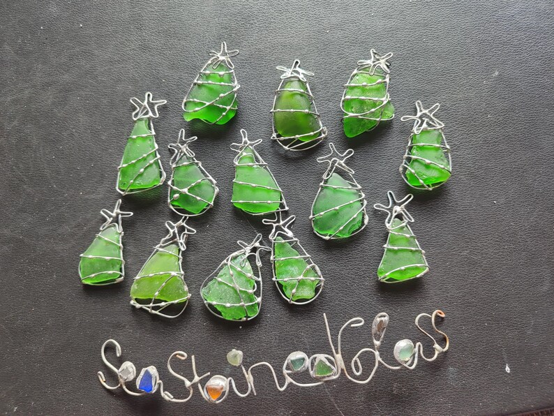 May include: Twelve green glass Christmas tree ornaments wrapped in silver wire. The ornaments are arranged in a semi-circle on a gray surface. The text "Sea Stained Glass" is written in silver wire below the ornaments.