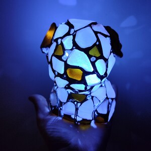 May include: A white and yellow stained glass dog figurine with black accents. The figurine is illuminated from within, casting a blue glow.