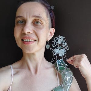 Puede incluir: Una figura decorativa de sirena hecha con alambre de tono plateado y diversas piezas de vidrio marino en tonos verdes y blancos esmerilados. La sirena presenta una cabeza y torso esculpidos en alambre y una cola construida con mosaico de vidrio.