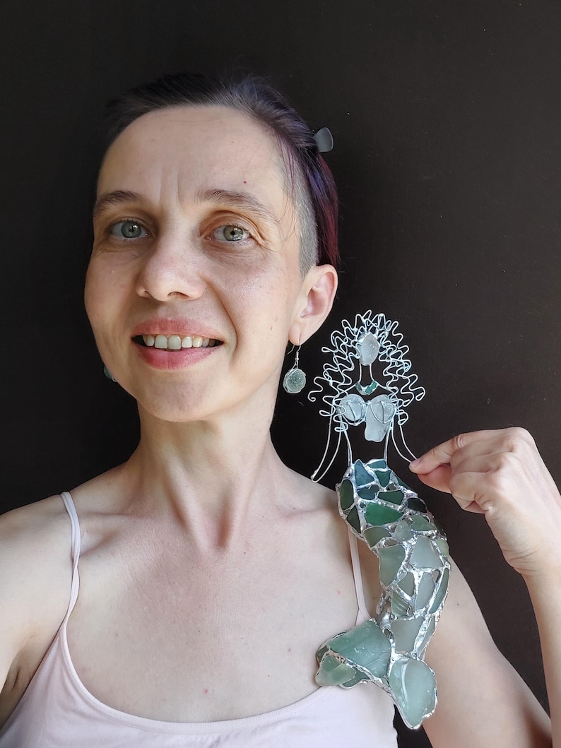 Puede incluir: Una figura decorativa de sirena hecha con alambre de tono plateado y diversas piezas de vidrio marino en tonos verdes y blancos esmerilados. La sirena presenta una cabeza y torso esculpidos en alambre y una cola construida con mosaico de vidrio.
