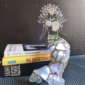 Puede incluir: Una escultura de sirena hecha de alambre plateado y piezas de vidrio marino iridiscente. La figura presenta una cabeza y un torso formados por alambre con una cola de mosaico. Est&aacute; colocada frente a tres libros apilados titulados L'Adversaire, The Queer Evangelist y Salman Rushdie.
