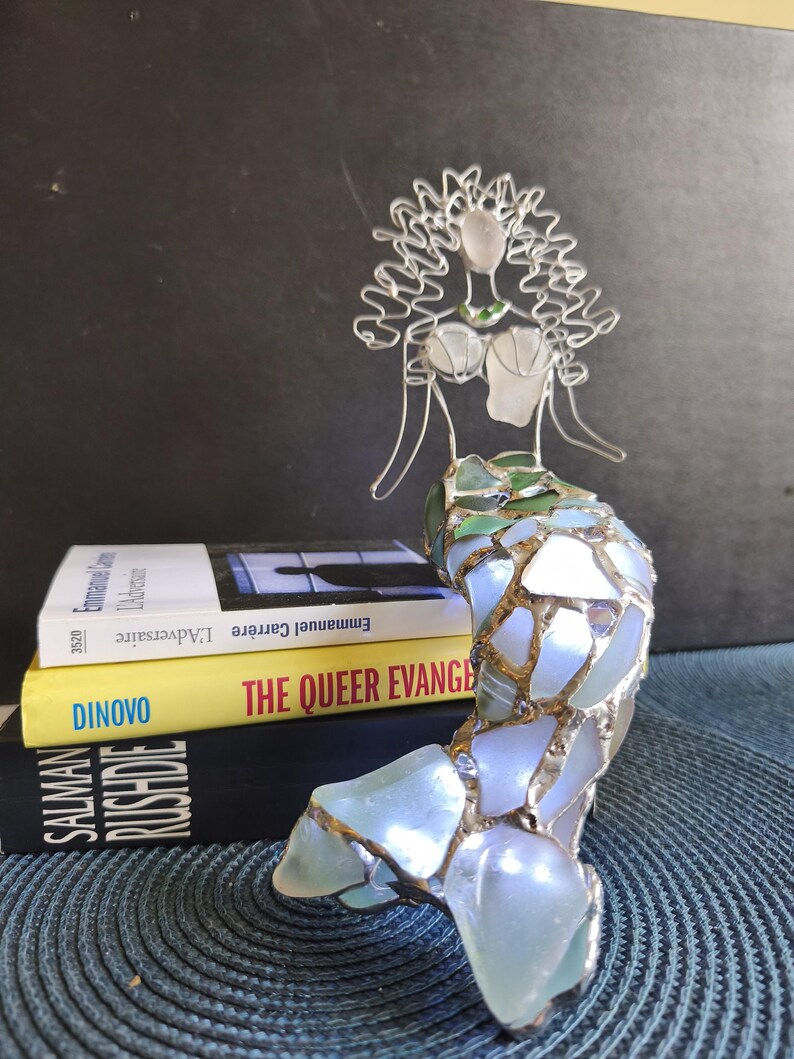 Puede incluir: Una escultura de sirena hecha de alambre plateado y piezas de vidrio marino iridiscente. La figura presenta una cabeza y un torso formados por alambre con una cola de mosaico. Est&aacute; colocada frente a tres libros apilados titulados L'Adversaire, The Queer Evangelist y Salman Rushdie.