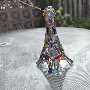 May include: A stained glass Eiffel Tower ornament in a variety of colours. The miniature tower is made of different coloured glass pieces, including red, blue, green, and yellow, held together with silver solder. A silver string is attached for hanging.
