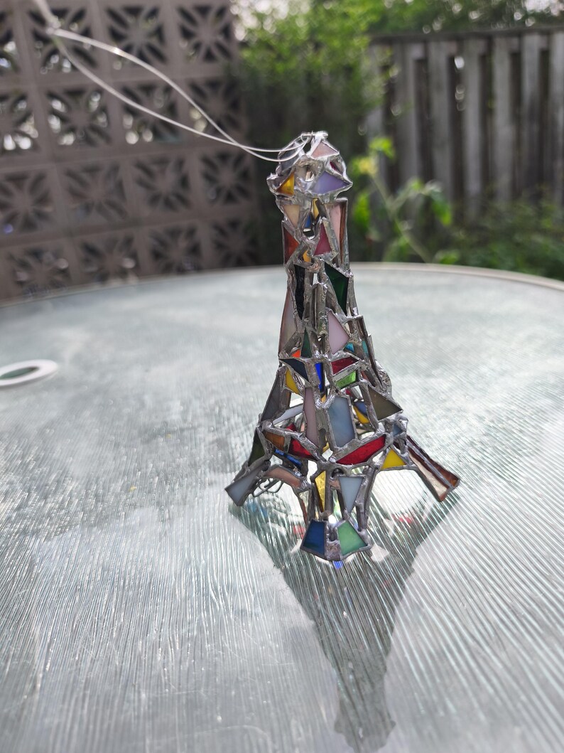 May include: A stained glass Eiffel Tower ornament in a variety of colours. The miniature tower is made of different coloured glass pieces, including red, blue, green, and yellow, held together with silver solder. A silver string is attached for hanging.