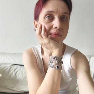 May include: Three silver bracelets with gemstone accents. The bracelets are stacked on top of each other. The gemstones are purple and blue. The bracelets have a wire design.