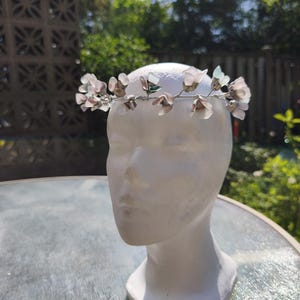 May include: A delicate floral crown featuring light pink and green flower accents. The crown is made of wire and is displayed on a white mannequin head. The background includes greenery and a sunny sky, suggesting outdoor use.