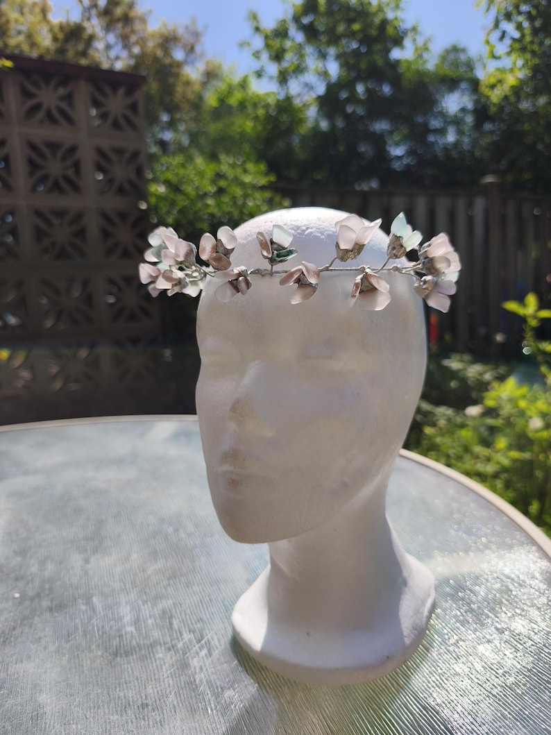 May include: A delicate floral crown featuring light pink and green flower accents. The crown is made of wire and is displayed on a white mannequin head. The background includes greenery and a sunny sky, suggesting outdoor use.