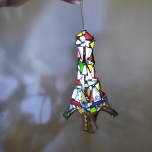 May include: A stained glass Eiffel Tower ornament, featuring a mosaic of coloured glass pieces. The ornament, suspended by a thin string, has a vibrant design with red, yellow, green, and blue hues. The design is intricate and detailed.