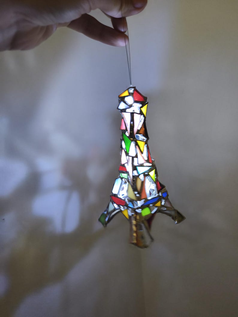 May include: A stained glass Eiffel Tower ornament, featuring a mosaic of coloured glass pieces. The ornament, suspended by a thin string, has a vibrant design with red, yellow, green, and blue hues. The design is intricate and detailed.
