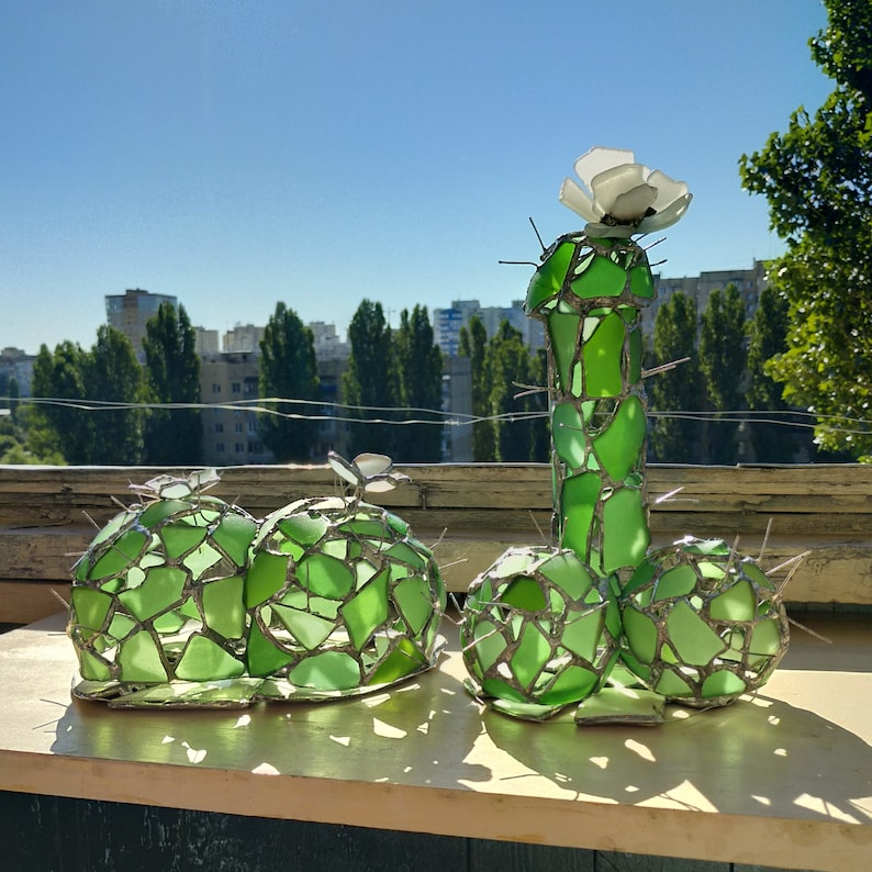 May include: A green glass sculpture of a cactus with a white flower on top. The sculpture is made of many small pieces of glass that are joined together.