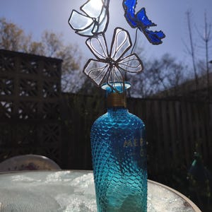 May include: A turquoise glass bottle with a textured surface, topped with a decorative glass flower arrangement. The flowers are in shades of blue and clear glass. The bottle is on a glass table, with a bright sun in the background.