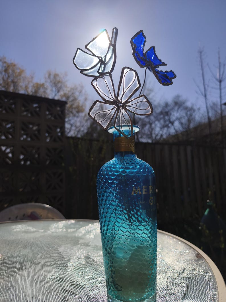 May include: A turquoise glass bottle with a textured surface, topped with a decorative glass flower arrangement. The flowers are in shades of blue and clear glass. The bottle is on a glass table, with a bright sun in the background.