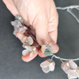 May include: A delicate floral crown featuring clusters of translucent, petal-shaped beads in shades of pink, peach, and green. The beads are attached to a thin, silver-colored wire, creating a whimsical and elegant accessory. The crown is held by a hand.