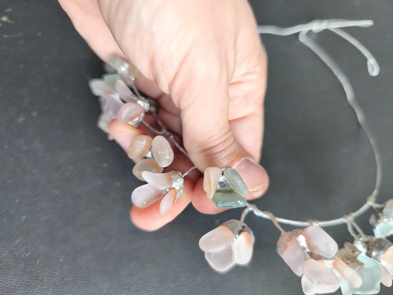May include: A delicate floral crown featuring clusters of translucent, petal-shaped beads in shades of pink, peach, and green. The beads are attached to a thin, silver-colored wire, creating a whimsical and elegant accessory. The crown is held by a hand.