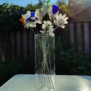 Op de afbeelding: Een boeket van zeeglasbloemen in een heldere glazen vaas. De bloemen zijn gemaakt van verschillende kleuren zeeglas, waaronder wit, blauw, groen en oranje. De vaas staat op een witte tafel met de woorden "Sea Stained Glass" in zwart draad.