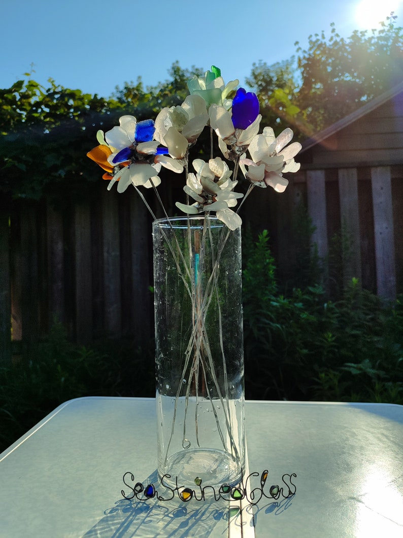 Op de afbeelding: Een boeket van zeeglasbloemen in een heldere glazen vaas. De bloemen zijn gemaakt van verschillende kleuren zeeglas, waaronder wit, blauw, groen en oranje. De vaas staat op een witte tafel met de woorden "Sea Stained Glass" in zwart draad.