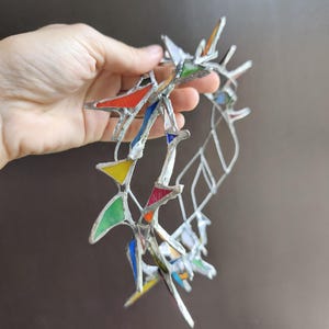 May include: A circular crown constructed from silver-toned metal solder and small, multi-colored stained glass shards in triangular shapes. The glass pieces feature vibrant red, yellow, green, blue, and orange hues, arranged in a jagged, open-work pattern.