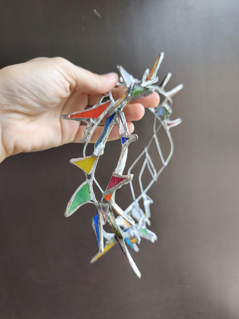 May include: A circular crown constructed from silver-toned metal solder and small, multi-colored stained glass shards in triangular shapes. The glass pieces feature vibrant red, yellow, green, blue, and orange hues, arranged in a jagged, open-work pattern.