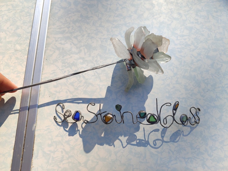 Op de afbeelding: Een witte zeeglasbloem met een draadsteel en de woorden "Sea Stained Glass" geschreven in draad met zeeglasaccenten.