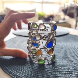 May include: A silver metal candle holder with colorful glass inserts. The candle holder has a cylindrical shape with multiple tiers, each adorned with various colored glass pieces, including blue, green, yellow, and clear. The design is intricate and decorative.
