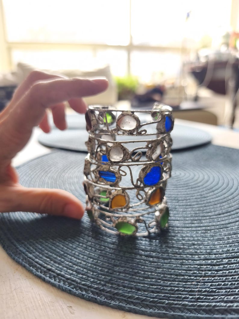 May include: A silver metal candle holder with colorful glass inserts. The candle holder has a cylindrical shape with multiple tiers, each adorned with various colored glass pieces, including blue, green, yellow, and clear. The design is intricate and decorative.