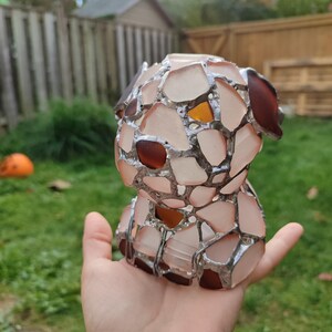 May include: A small, hand-crafted glass sculpture of a dog. The sculpture is made of many small pieces of glass, which are fused together with silver solder. The glass is a mix of pink, brown, and clear.