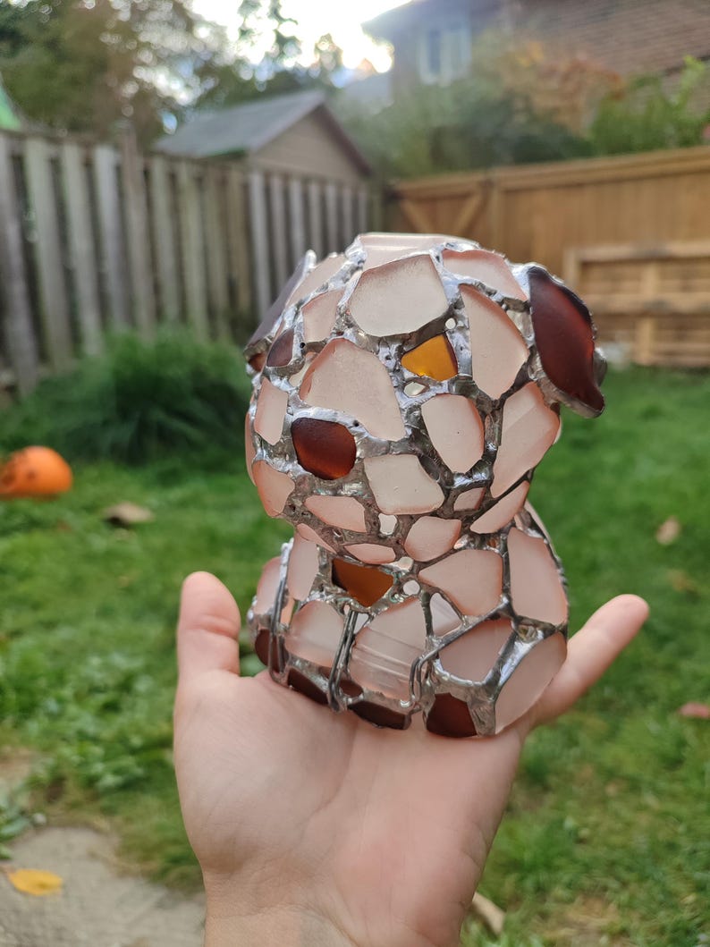 May include: A small, hand-crafted glass sculpture of a dog. The sculpture is made of many small pieces of glass, which are fused together with silver solder. The glass is a mix of pink, brown, and clear.