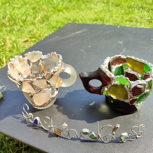 May include: Two small cups made of stained glass, one with pink and orange glass pieces and the other with green, yellow, and brown glass pieces. The cups are sitting on a black surface with the words "Sea Glass" written in silver wire.