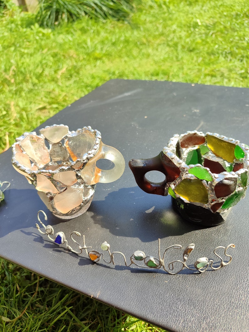 May include: Two small cups made of stained glass, one with pink and orange glass pieces and the other with green, yellow, and brown glass pieces. The cups are sitting on a black surface with the words "Sea Glass" written in silver wire.