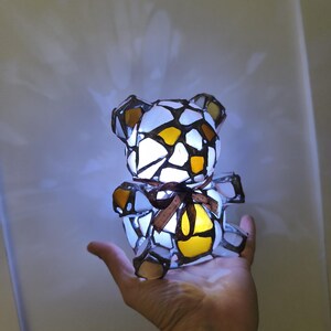 May include: A stained glass teddy bear lamp with a brown ribbon around its neck. The bear is made of white, yellow, and brown glass pieces, and it is lit from within. The lamp is held in a person's hand.