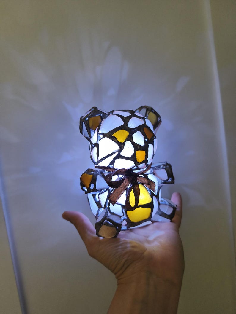 May include: A stained glass teddy bear lamp with a brown ribbon around its neck. The bear is made of white, yellow, and brown glass pieces, and it is lit from within. The lamp is held in a person's hand.
