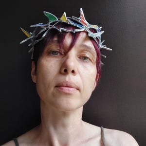 May include: A crown featuring a silver-toned metal frame with sharp, pointed edges and small, multi-colored stained glass inserts in shades of red, yellow, green, and blue. The structure resembles a crown of thorns with a metallic finish.
