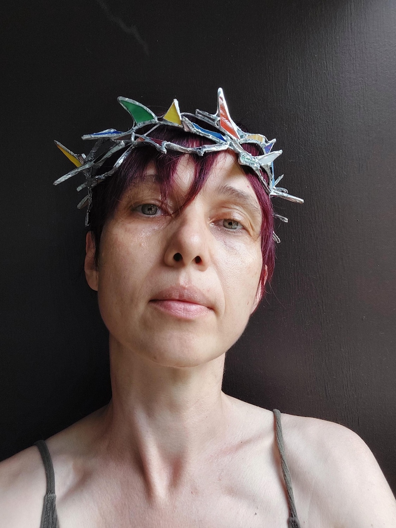 May include: A crown featuring a silver-toned metal frame with sharp, pointed edges and small, multi-colored stained glass inserts in shades of red, yellow, green, and blue. The structure resembles a crown of thorns with a metallic finish.