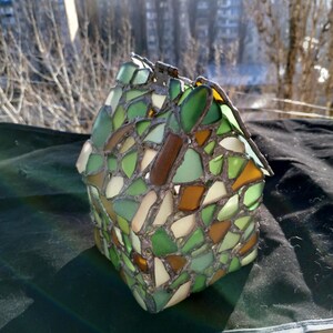 May include: A small house-shaped lamp made of stained glass with green, brown, and white pieces. The lamp is sitting on a dark green surface.