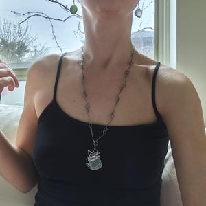 May include: A silver-toned necklace with a cat pendant, paired with matching earrings. The cat pendant is a wireframe design. The model is wearing a black tank top. The earrings are round and green.