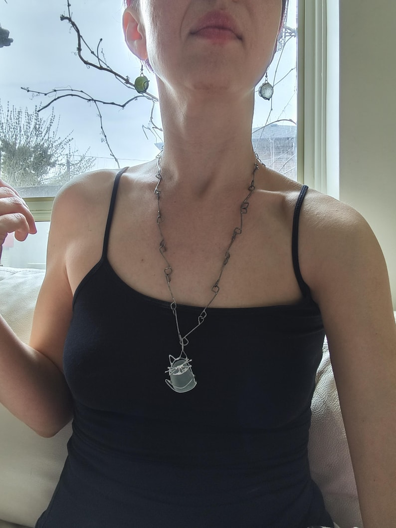 May include: A silver-toned necklace with a cat pendant, paired with matching earrings. The cat pendant is a wireframe design. The model is wearing a black tank top. The earrings are round and green.