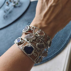 May include: Silver-toned metal bracelets with irregularly shaped, colorful stones. The bracelets are worn on a wrist, showcasing the unique design and textures of the stones. The stones are various colors, including blue, pink, and gray.