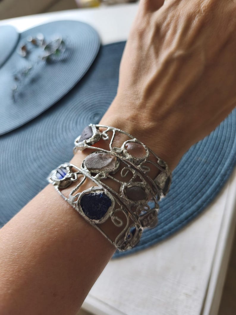 May include: Silver-toned metal bracelets with irregularly shaped, colorful stones. The bracelets are worn on a wrist, showcasing the unique design and textures of the stones. The stones are various colors, including blue, pink, and gray.