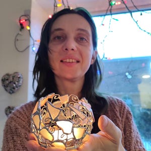 May include: A woman holds a white and silver candle holder made of glass and metal. The candle holder is shaped like a sphere and has a delicate, intricate design. The candle holder is lit and the flame is visible.