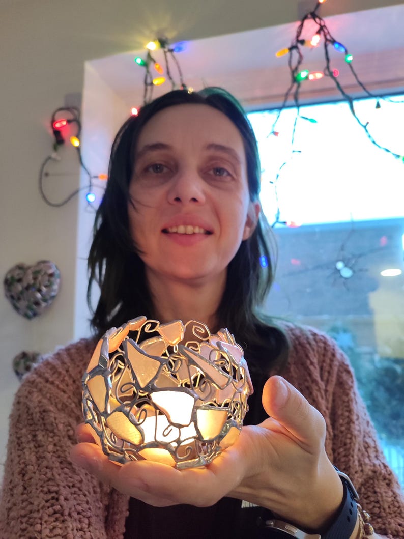 May include: A woman holds a white and silver candle holder made of glass and metal. The candle holder is shaped like a sphere and has a delicate, intricate design. The candle holder is lit and the flame is visible.