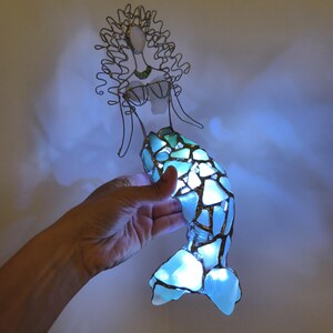 Puede incluir: Una escultura de sirena hecha de alambre y piezas de vidrio marino azul claro. La cola est&aacute; iluminada desde el interior, proyectando un brillo intenso a trav&eacute;s de los segmentos de vidrio transl&uacute;cido.