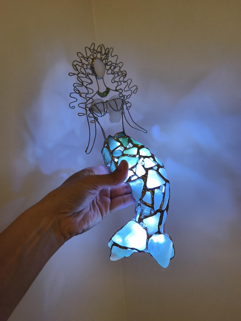 Puede incluir: Una escultura de sirena hecha de alambre y piezas de vidrio marino azul claro. La cola est&aacute; iluminada desde el interior, proyectando un brillo intenso a trav&eacute;s de los segmentos de vidrio transl&uacute;cido.