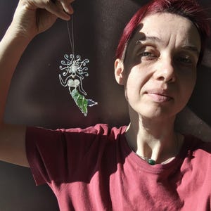 Puede incluir: Adorno de sirena hecho a mano con un marco de alambre plateado, un busto blanco y una cola verde. El cabello de la sirena es un dise&ntilde;o arremolinado. El adorno est&aacute; suspendido por un hilo fino. El fondo es una superficie oscura y texturizada.