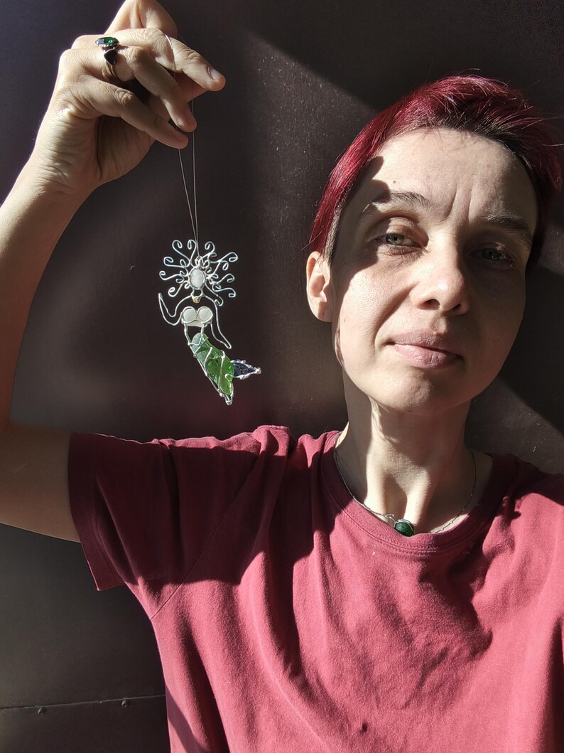 Puede incluir: Adorno de sirena hecho a mano con un marco de alambre plateado, un busto blanco y una cola verde. El cabello de la sirena es un dise&ntilde;o arremolinado. El adorno est&aacute; suspendido por un hilo fino. El fondo es una superficie oscura y texturizada.