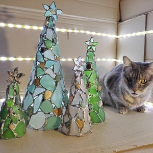 Pode incluir: Cinco &aacute;rvores de Natal decorativas de vidro em v&aacute;rios tamanhos e tons de verde, azul e branco. Cada &aacute;rvore &eacute; adornada com uma estrela no topo. Um gato cinza malhado est&aacute; por perto. As &aacute;rvores s&atilde;o iluminadas por uma fileira de luzes.