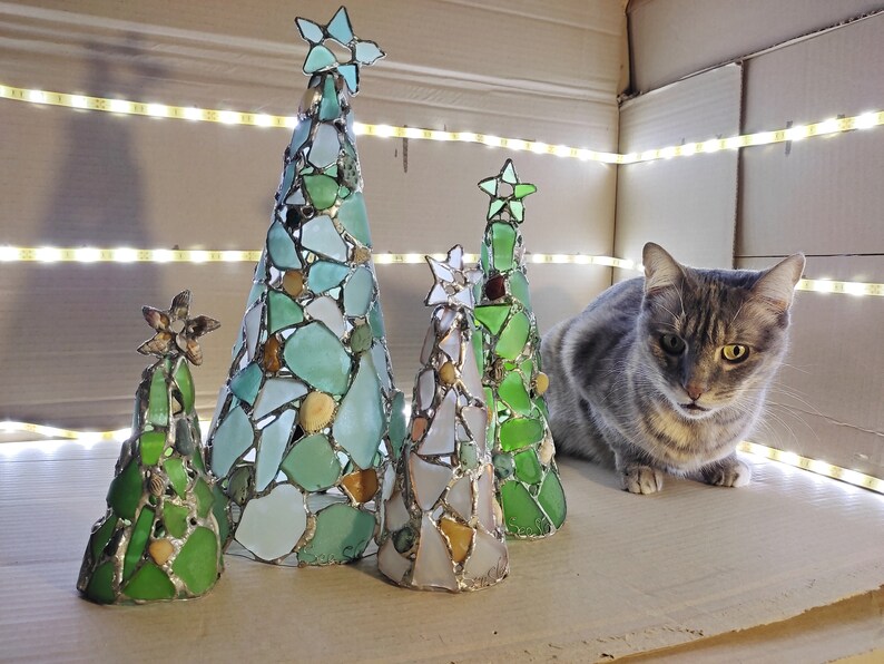 Pode incluir: Cinco &aacute;rvores de Natal decorativas de vidro em v&aacute;rios tamanhos e tons de verde, azul e branco. Cada &aacute;rvore &eacute; adornada com uma estrela no topo. Um gato cinza malhado est&aacute; por perto. As &aacute;rvores s&atilde;o iluminadas por uma fileira de luzes.